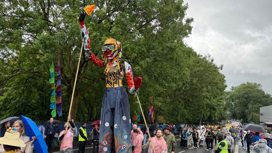 A largescale puppet held up by sticks carried by men in clear ponchos over pink tops. The puppet appears to be female and is yellow, red and blue, wearing goggles and smiling. A number of people can be see walking behind in the centre of a rainy street, with others watching from the pavements. Many are carrying umbrellas.
