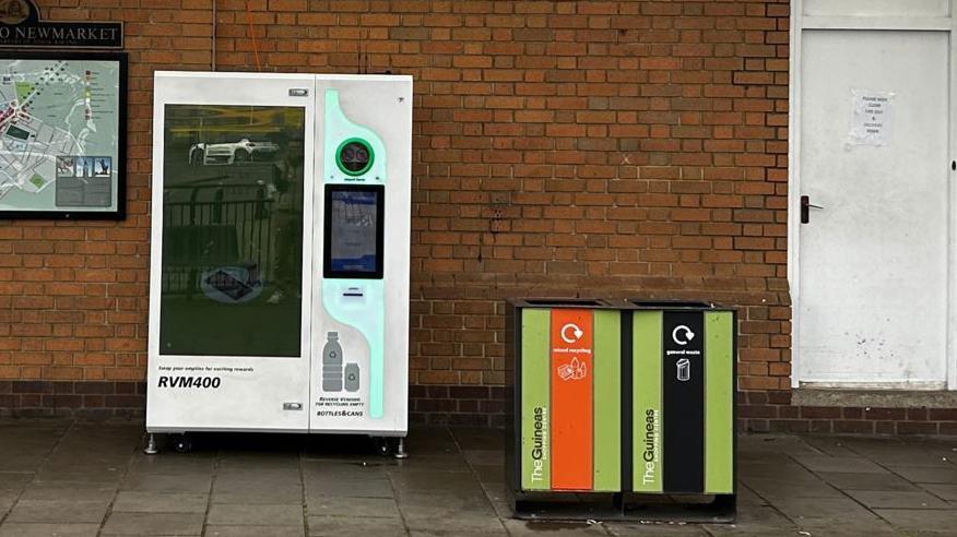 A picture of the reverse vending machine which is a white long box featuring two digital screens and a circular hole to deposit bottles in. 