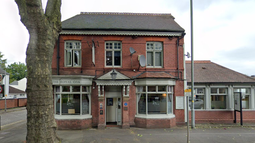 The Royal Oak Pub has a large tree on the pavement outside it. it is a red brick pub with grey/green panels around its windows and entrance way.