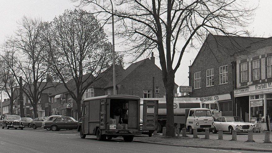 A black and white image of a van with open back doors, a tanker lorry and a row of shops near a road