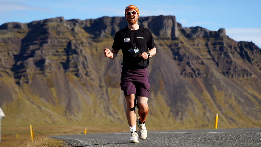 A man wearing a black t-shirt, purple shorts, an orange hat and a pair of sunglasses running on a flat, empty road with mountains behind him. He's running and smiling at the camera.