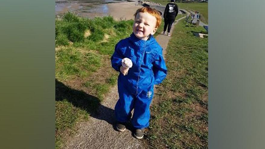 George Herd is smiling as he holds an ice cream cone while standing on a path by the sea. He has red hair and is wearing a blue all-in-one waterproof suit.