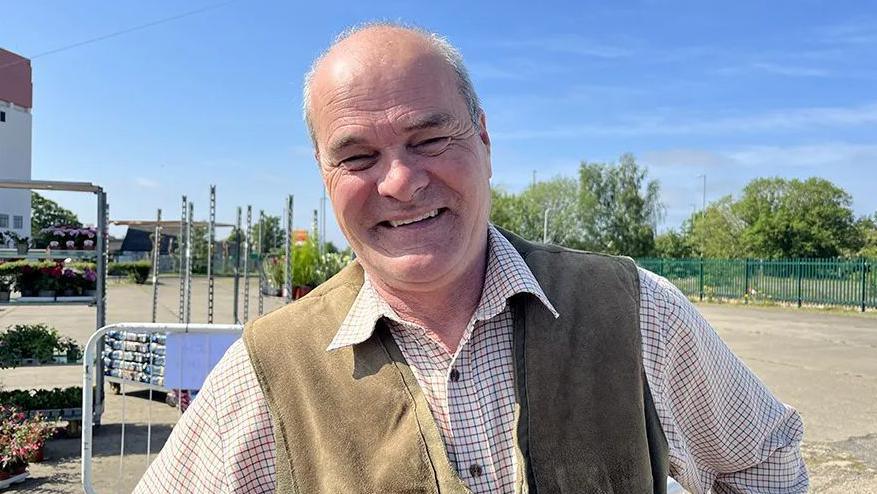 Stephen Timewell is smiling directly at the camera. He is wearing a checked shirt and brown waistcoat. In the background, flowers and plants can be seen stacked up on trolleys. The sky is a bright blue. 