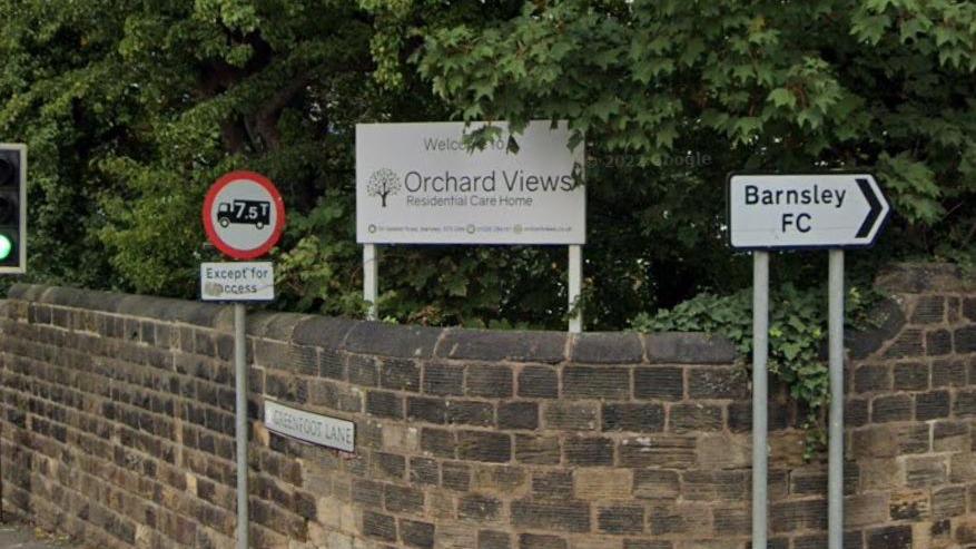 An image of the corner of Greenfoot Lane in Gawber, Barnsley. There is a chest-high stone wall.  Behind it is a sign saying "Welcome to Orchard Views care home".  In front of the wall are two road signs.  he sign to the left bans lorries over 7.5 tonnes, the sign to the right of the picture directs to Barnsley FC.