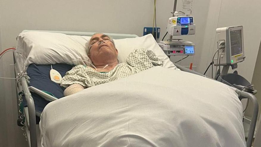 Paul lies asleep in a hospital bed, the bed is propped up at an angle and has multiple machines around him. He is covered by a white blanket