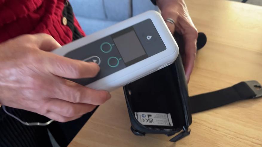 The pump device is white, with a grey front, featuring a screen and buttons. Mrs Fuller is inserting into a black bag which has straps to affix the device to her midriff. She is holding the device in her right hand, and the bag in her left. The worktop is wood-effect and she is wearing a red knitted jersey and black trousers.