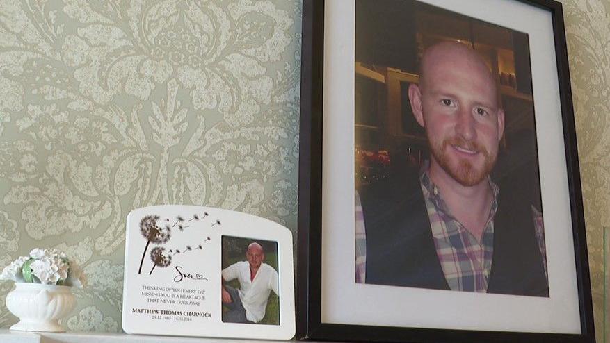 A small white vase containing white flowers is next to two photo frames with pictures of Matthew Charnock. One is a white frame with an image of two dandelion flowers and the words 'Son, thinking of you every day, missing you is a heartache that never goes away' with Mr Charnock's name and his birth date and the date that he died.