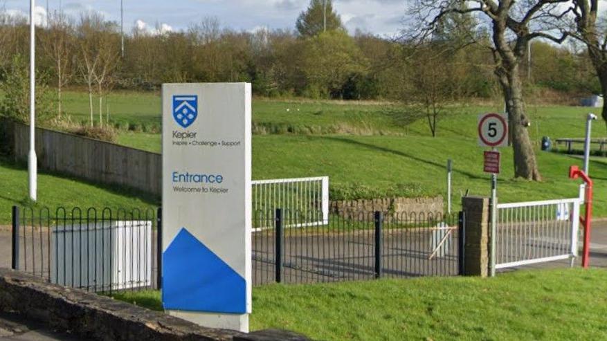 The blue and white entrance sign at the gates of Kepier Academy in Houghton-le-Spring. A red 5mph speed sign is near the school fences. There is grassland and trees nearby.