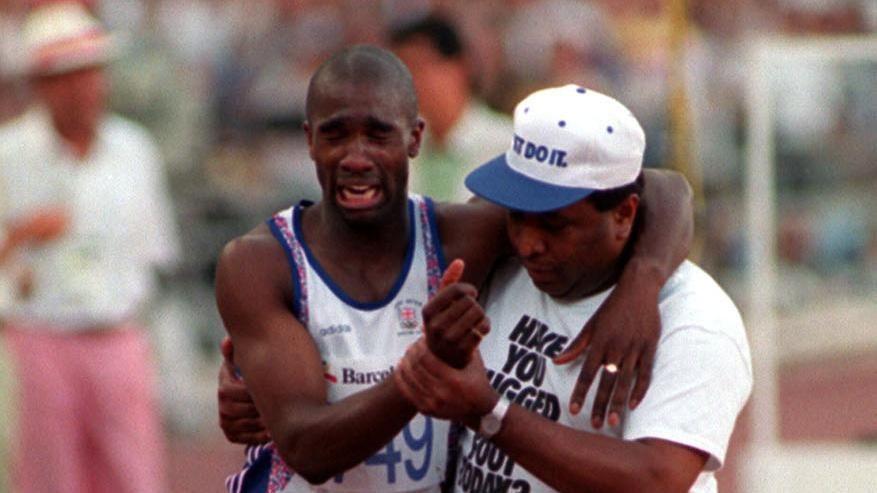 Derek Redmond in running gear being helped by his father who is wearing a blue and white cap