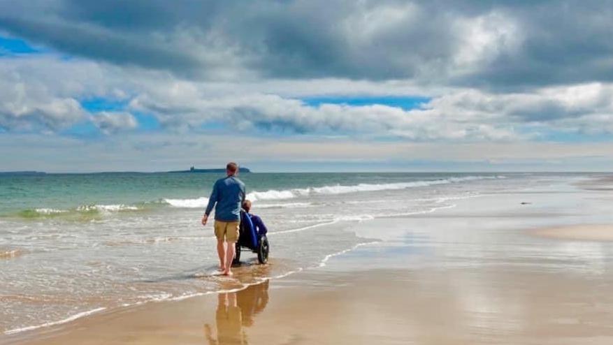 View of a sandy beach under cloudy but bright skies. A man, wearing khaki shorts and a blue top, is walking on the shoreline pushing someone in a wheelchair.