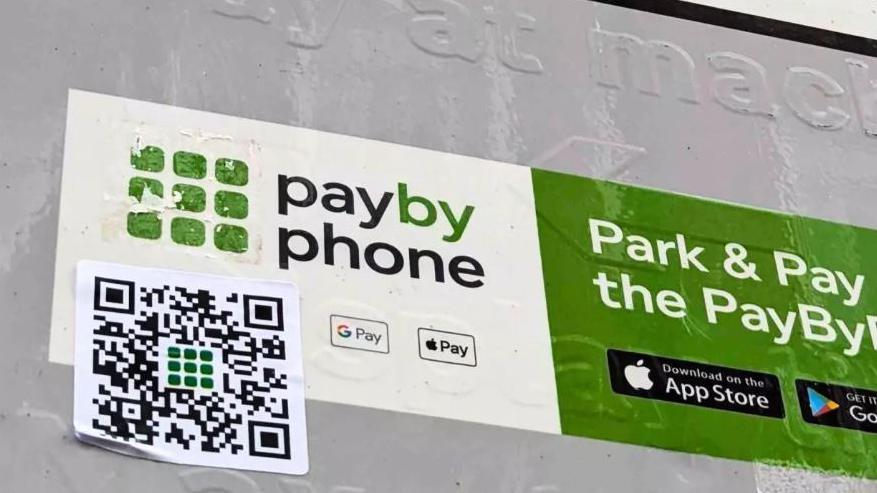 A white and black QR code sticker sits on a car parking payment sign. The sticker is crumpled on the bottom left corner.