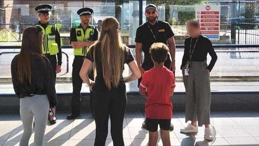 Picture shows the backs of three children, two girls wearing black tops and a boy wearing a red top, stood in front of two police officers who are to the left of the picture at the back and two other people to the right of the police officers, one wearing a black top and baseball cap.