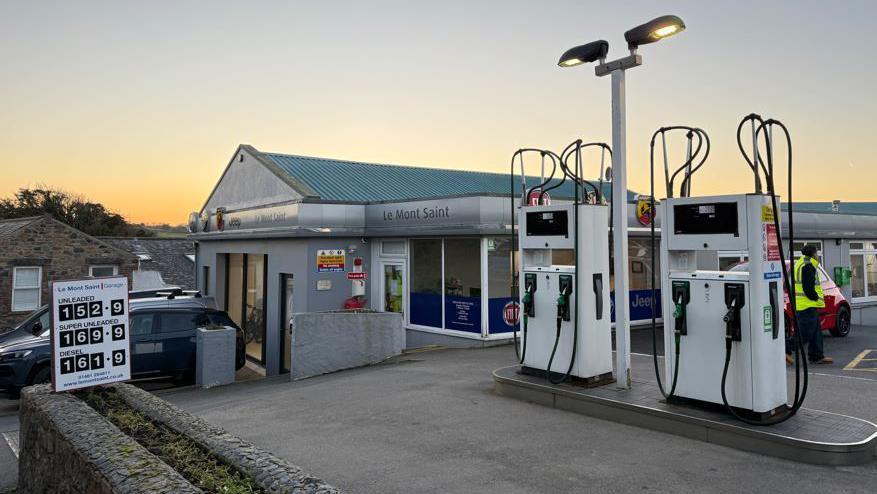 The Le Mont Saint garage. It is a silver building with cars outside it. There are two fuel pumps and a fuel price board. A person is walking across the forecourt in the background as the sun sets.