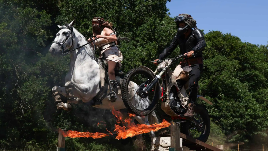A person with a white skirt and top on, with long hair and a mask on their face is riding a white horse which is jumping in the air. Next to it, a motorbike rider is doing a jump on a black bike off a ramp with flames next to it.