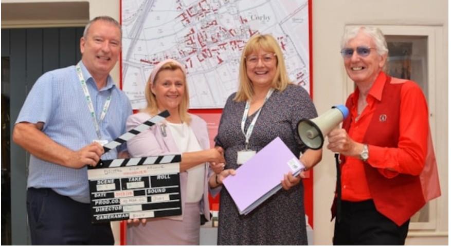 A man in a blue shirt holds a film take board next to a woman in a pink dress, a woman in a black floral dress and a man in a red blazer holding a microphone.