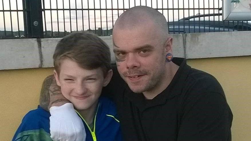 Jamie Jennings as a young boy with Brian McMaster who is looking into the hammer. Jamie has short hair and is smiling with his mouth closed as Brian has his arm over Jamie's shoulder for the photo. Brian, who is bald, has a large tattoo on the left side of his face that goes around his eye.