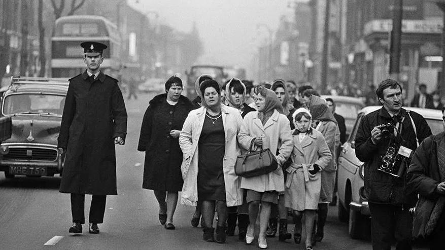 Lillian Bilocca leads a group of women through a busy London street. A police officer accompanies the women and there are a couple of press photographers in the picture too. 