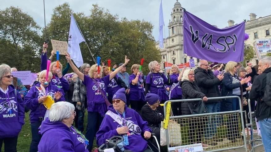 A large group of women in purple t-shirts and jumpers standing outside Parliament with banners and signs.