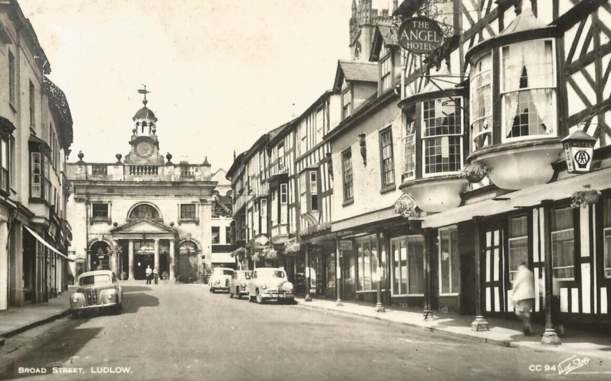 A black and white photo of a street with black and white timbered buildings down the right hand side and a building with a grand entrance with pillars at the end of the street