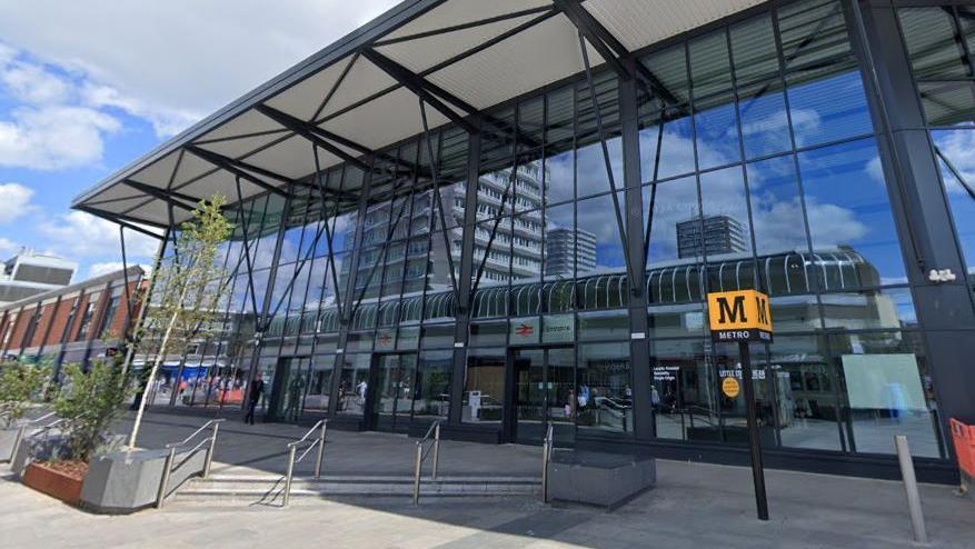 Streetview of Sunderland station. It is a large glass building. There is a black pole outside, atop of which stands a yellow cube with a large black M and the word Metro written on it.