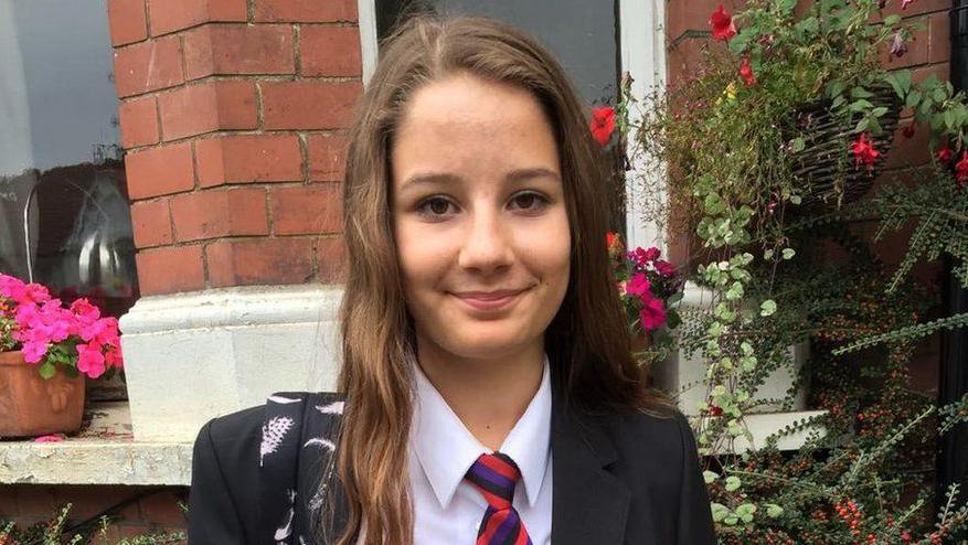 Molly Russell, shown wearing a school uniform and standing in front of a red-brick house. There are bright colourful flowers in pots on the window-ledge of the house behind her and foliage framing her face to the right of the image.
