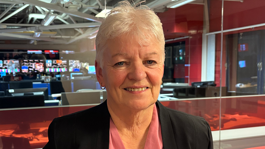 Bernie McNally is in a TV studio, she has short white hair and is wearing a black suit jacket and a pink top. She is smiling at the camera.