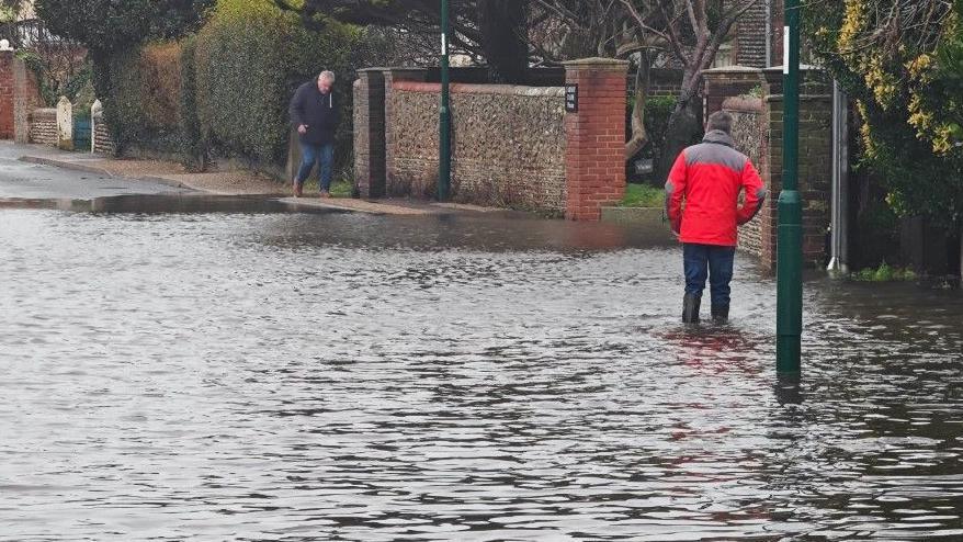 A man wading through water on a residential street. He is wearing a red jacket, jeans and green wellies and has his hands in his pockets.