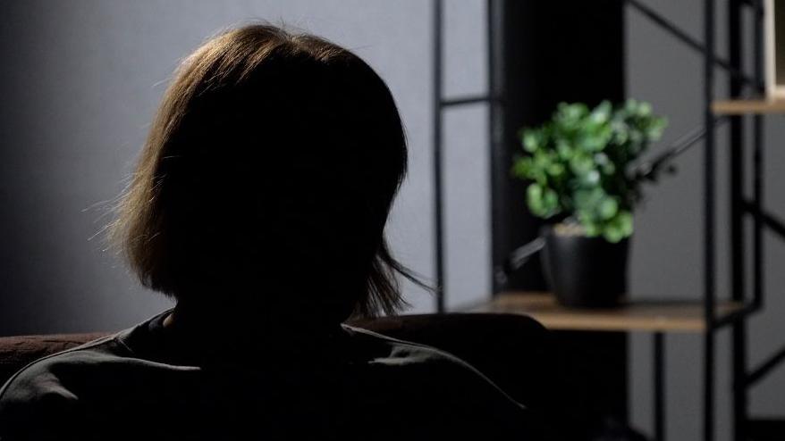 A woman sits in silhouette to protect the interviewee's anonymity. She is sitting in front of a grey background with plastic flowers on a wooden shelf. 