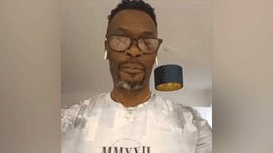 Calvin Olawale Babatunde is wearing a white t-shirt and dark rimmed glasses. He has a goatee which is grey at the bottom. 