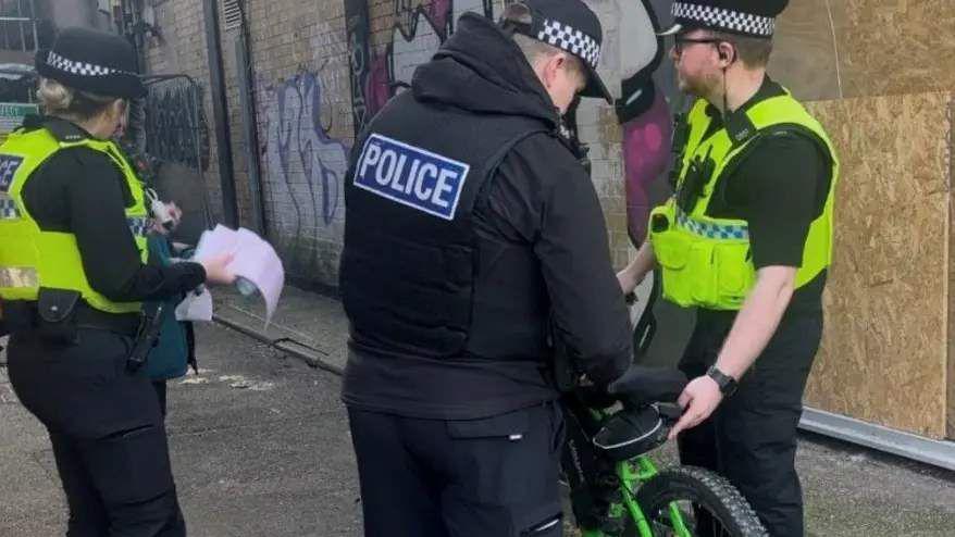 Three police officers are stood in a street with an e-bike