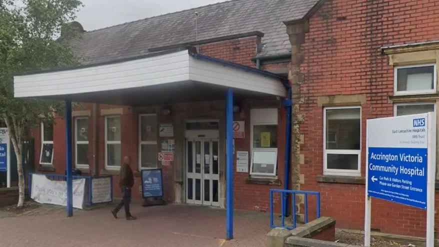 A street view image of Accrington Victoria Hospital, a red brick building with blue and white signs either side of a white awning with blue legs over the entrance and a man walking under it to the front door