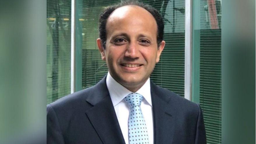 Prof Ahmed Ahmed smiling for the camera. He is wearing dark suit and a blue tie with black dots. He's standing outside in front of a window.