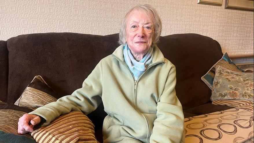 An elderly woman sits on a brown couch and smiles at the camera 