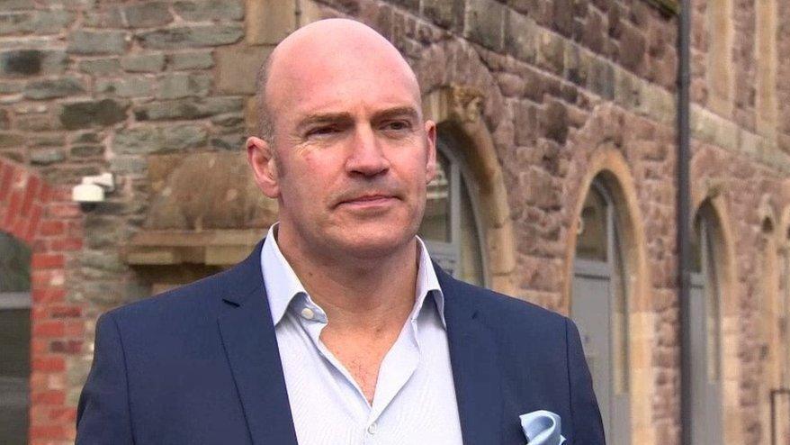 Steve Bradley is wearing a white shirt and blue jacket. He is standing in front of a red brick building.
