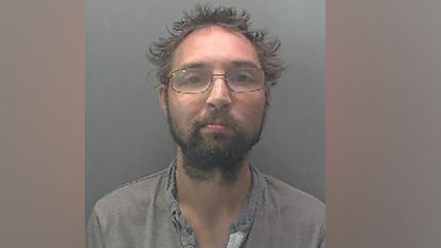 Police mugshot of a man with grey sticky-up hair and glasses. He is wearing a grey open-necked shirt