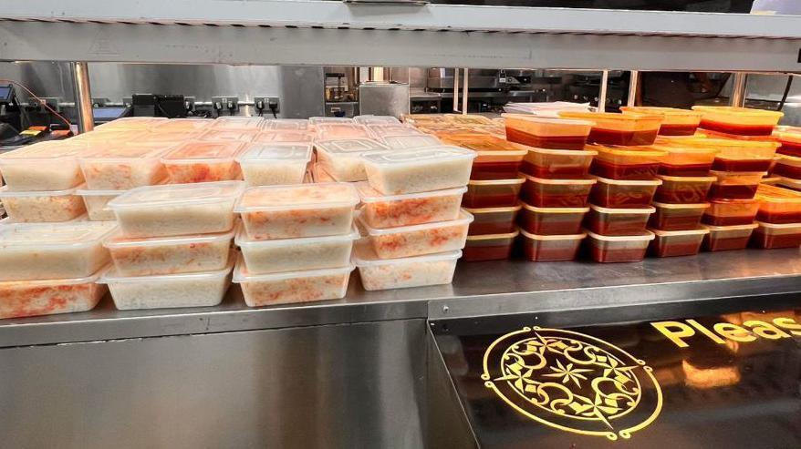 Dozens and dozens of tuppaware are stacked on a metal counter. The left half are filled with rice, the right half are filled with a red curry.