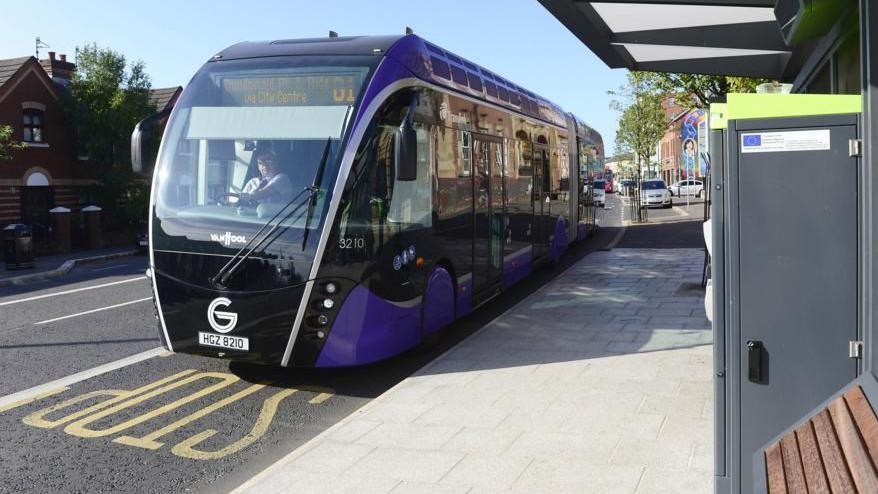 A large purple bus on a road, pavement and bus stop with ticket machine are visible on the right hand side