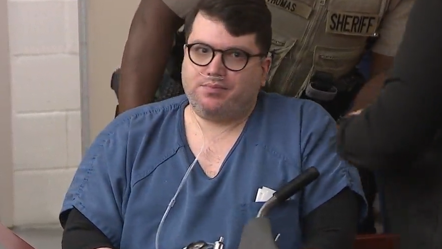 A clean-shaven Nicholas Rossi staring at the camera while sitting in court. in Utah. He is wearing loose-fitting prison scrubs and is carrying and attached to an oxygen tank