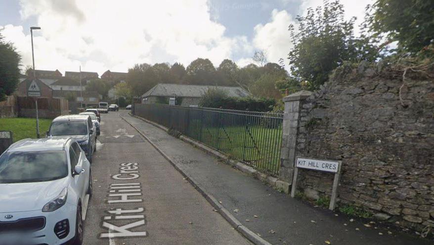 Google Street View image of Kit Hill Crescent in the Barne Barton area of Plymouth. Several vehicles are parked up on the side of the road. A small green area behind a set of iron railings is on the right hand side of the road.