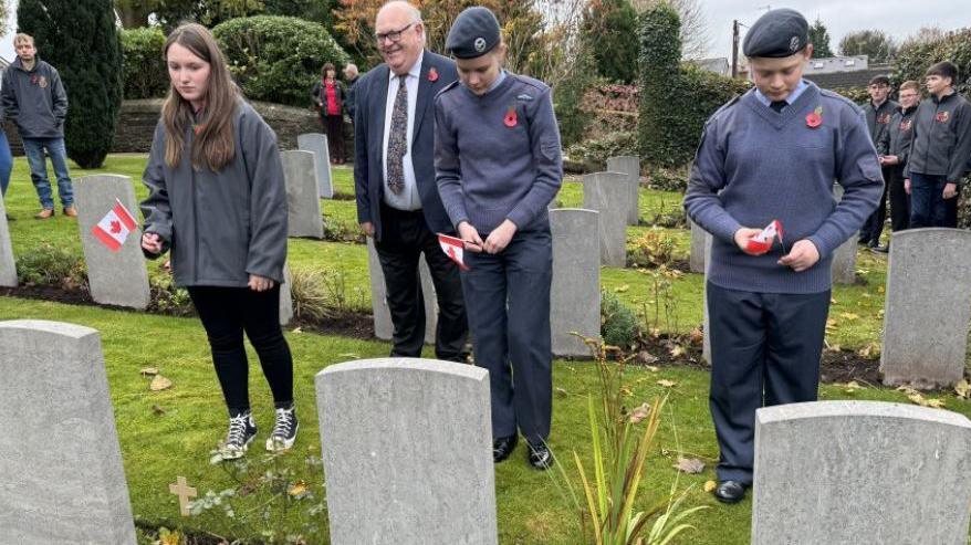 Three cadets holding Canadian flags in front of three war graves. There are onlookers in the background. Two are wearing blue cadet uniforms with berets, and one is wearing black leggings and a grey rain coat. 