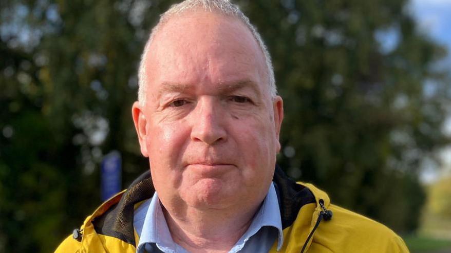 Close up headshot of middle aged white man with cropped grey hair, wearing a yellow rain coat and a blue shirt open at the neck 