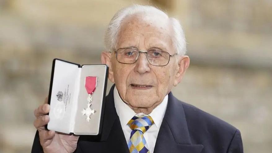 Harry Olmer holds up his MBE medal which is in a smart case. He wears a blue and gold chequered tie, a white shirt and navy suit jacket. He is in his 90s and has white hair and metal rimmed glasses.