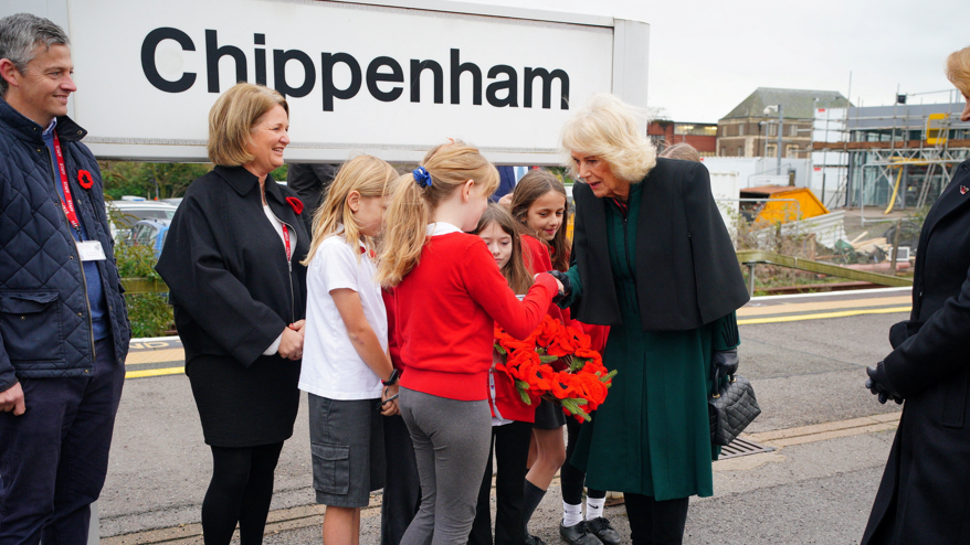 Queen Camilla wearing a green dress and holding a black bag shakes hands with a young girl holding a poppy wreath. A crowd of people surround them and a railway sign reads "Chippenham".