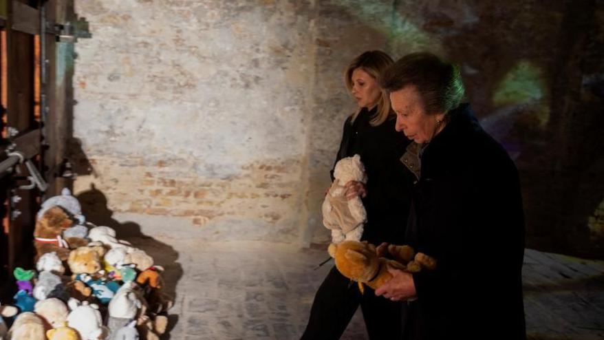 Two woman dressed in dark clothes solemnly approach a pile of soft toys on a darkened street.
