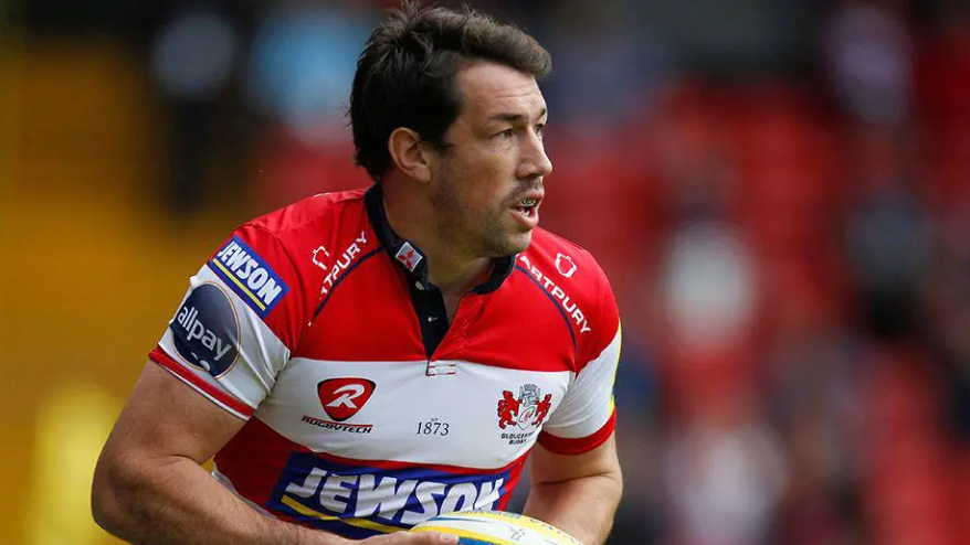 Tom Voyce running with a rugby ball during a professional match. He has dark hair and wears a mouth guard, and is looking away from the camera. He also wears a red and white Gloucester Rugby shirt. He is in focus, but the rest of the background is heavily blurred. 