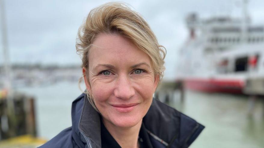 Fran Collins who has blonde hair and is wearing black coat standing on the quayside. Behind her is a red and white ferry.