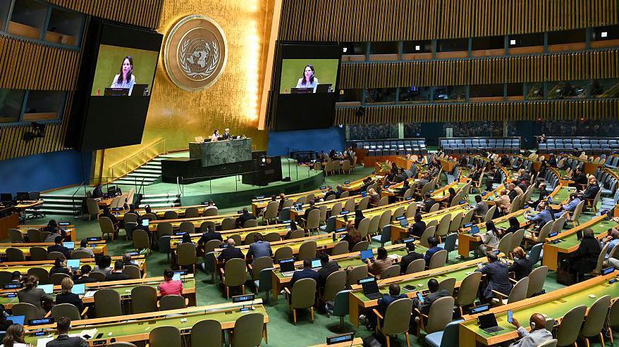 Inside the UN building rows of long desks and chairs face a stage and screens as participants watch a speaker join by video link.