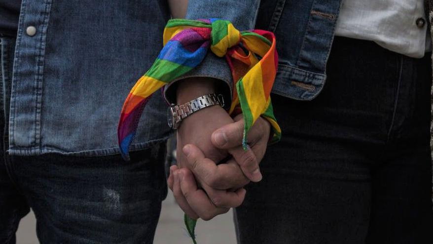 A same-sex couple, dressed in denim jackets, hold hands during an event to raise awareness of gay rights. Their wrists are tied together by a rainbow ribbon.