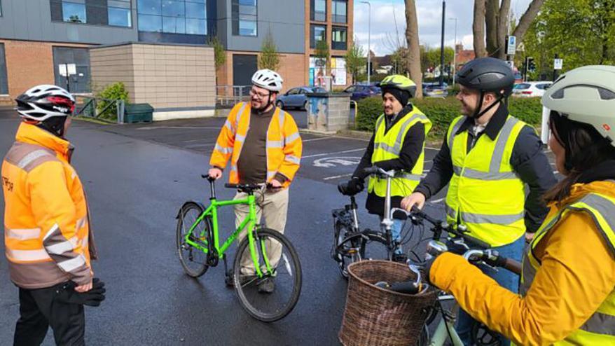 People on the training course wearing hi-visibility jackets and cycle helmets hold on to their bikes as they stand in a circle listening to an instructor