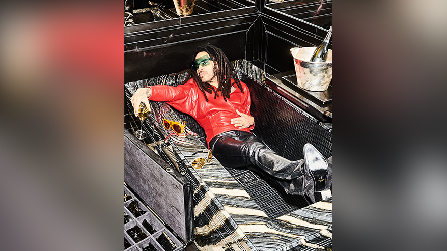 Lenny is lounging in an an empty black tiled bathtub. He has a glass of champagne in one hand and a bottle in a silver ice bucket beside the bath. He is wearing a red leather jacket and black leather trousers. He has dreadlocks and sunglasses.